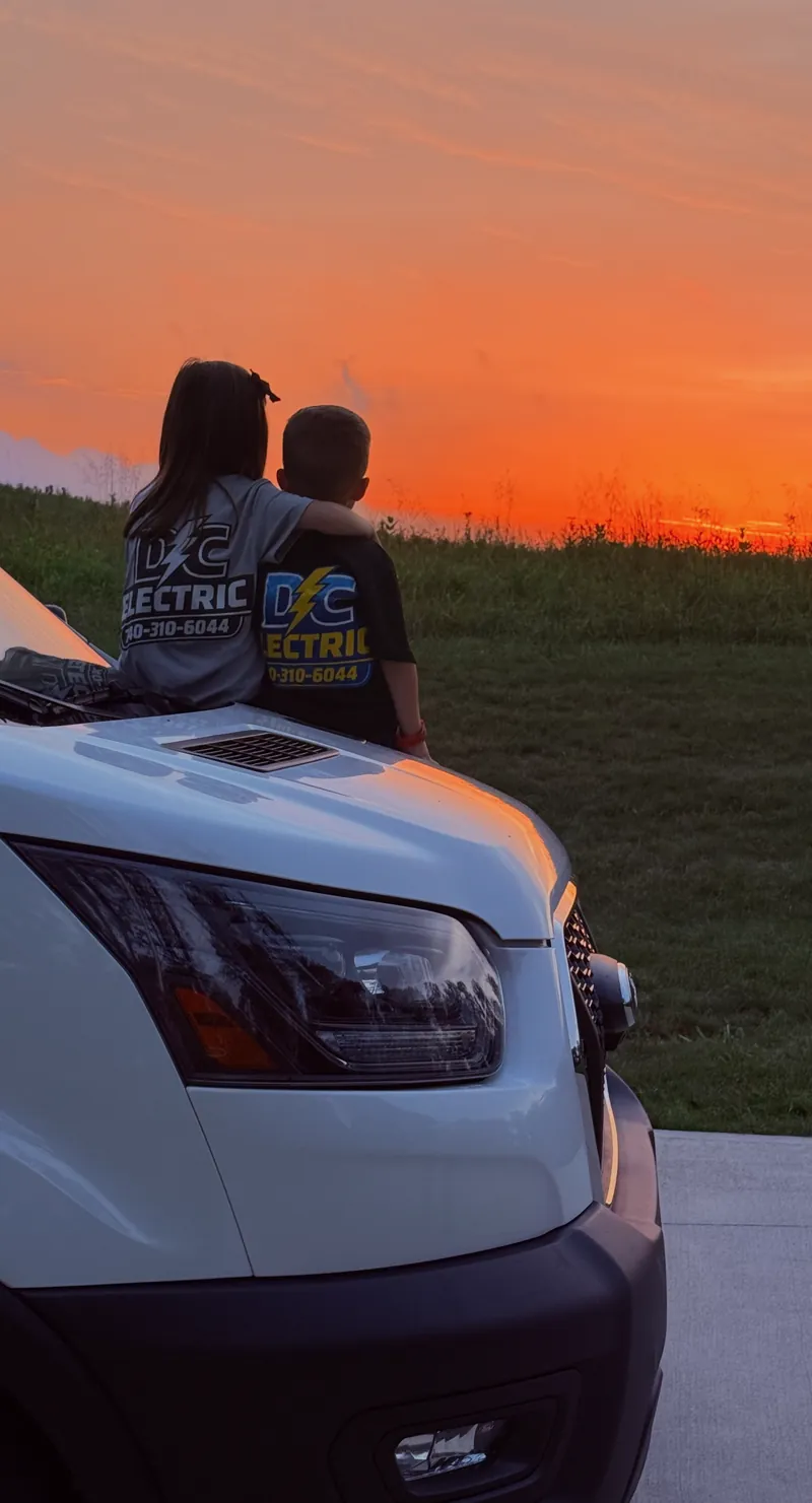 Two children in DC Electric shirts sitting on the hood of the work van, watching the sunset