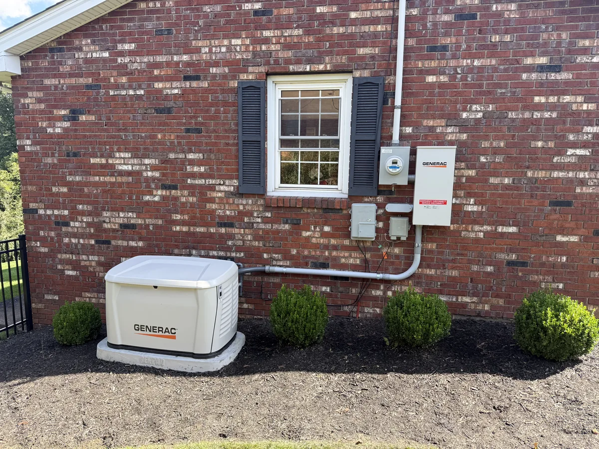 A Generac standby generator on its pad beside a brick house, with the meter, disconnect and transfer switch on the wall above the flowerbed, installed by DC Electric